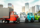 Electric rickshaws in various colors displayed on a city street with modern skyscrapers in the background.