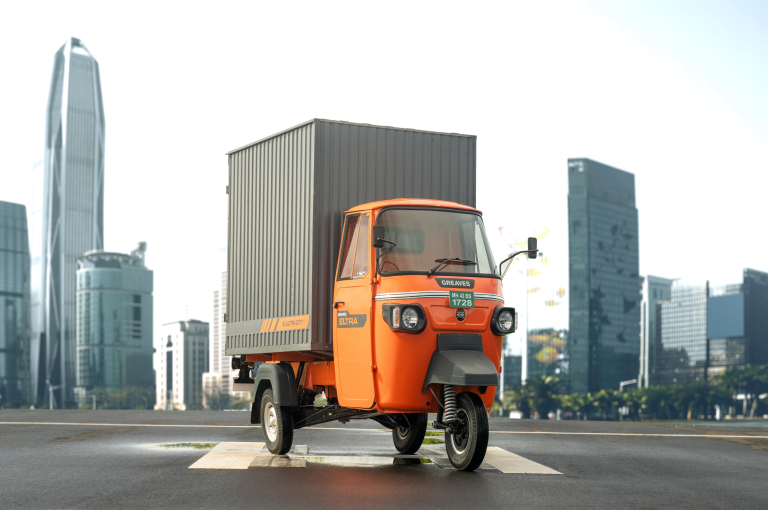 Electric three-wheeler cargo vehicle parked in an urban setting with skyscrapers in the background.