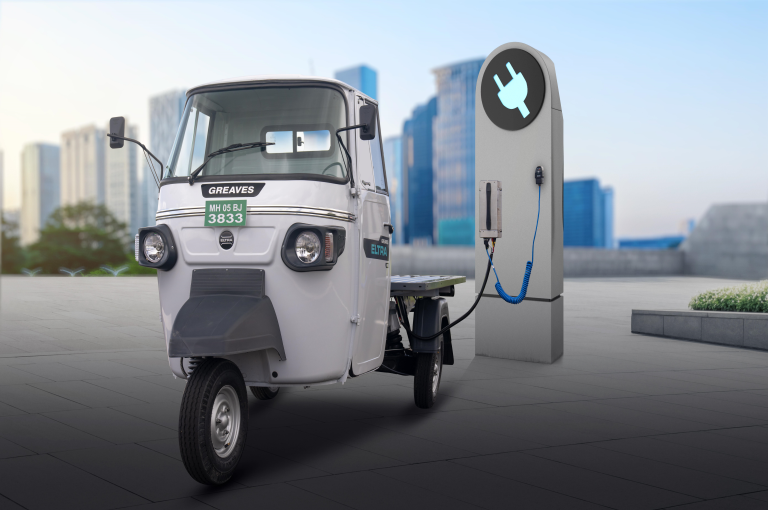 Electric three-wheeler charging at a charging station in an urban setting with skyscrapers in the background.